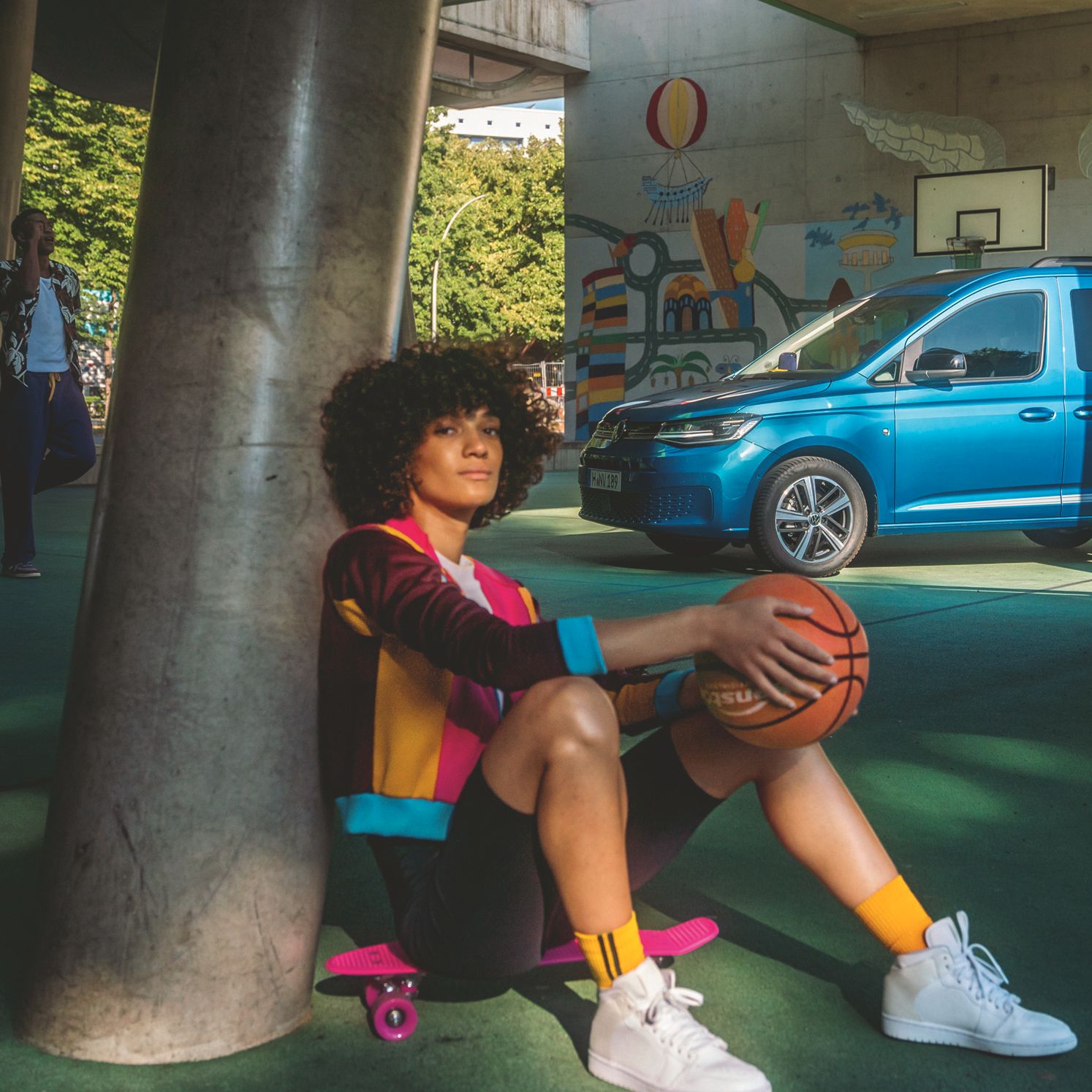 Ein blauer VW Caddy parkt auf einem Basketballplatz, eine Spielerin sitzt auf einem Skateboard davor