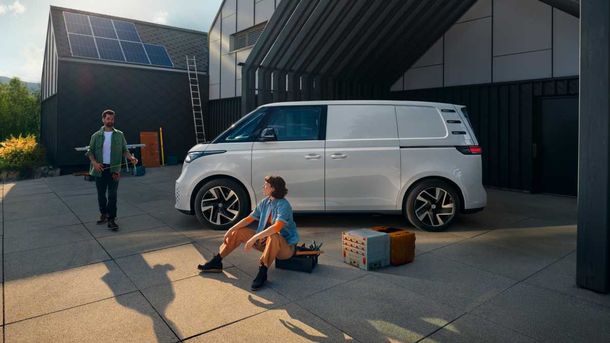 Ein weißer Volkswagen ID. Buzz Cargo vor einer Werkshalle, im Vordergrund sind ein Mann und eine Frau zu sehen
