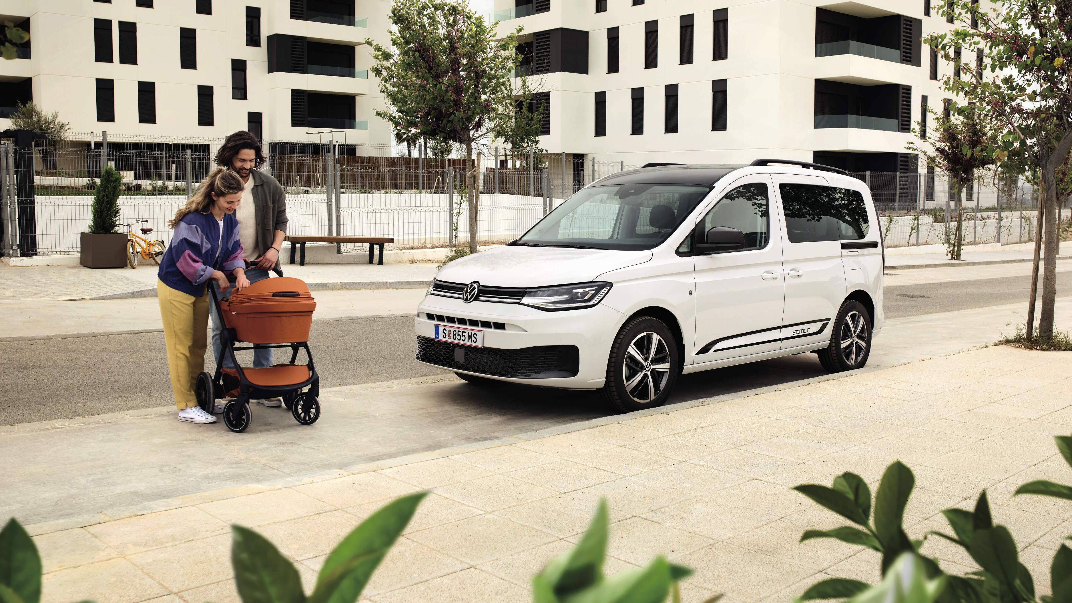 Zwei Erwachsene schieben ein Baby in einem Kinderwagen, Caddy Edition steht daneben