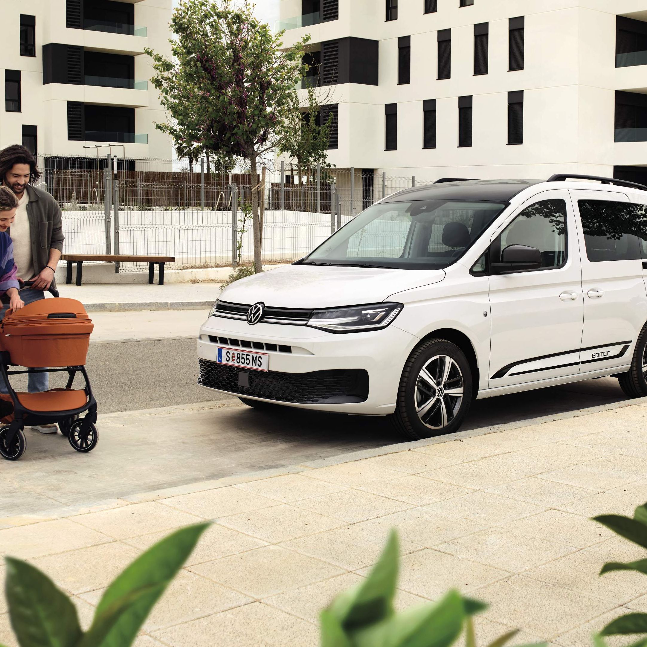 Zwei Erwachsene schieben ein Baby in einem Kinderwagen, Caddy Edition steht daneben